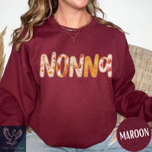 May include: Maroon sweatshirt with the word "NONNA" in a decorative, fall-themed font. The letters feature floral and plaid patterns in orange, yellow, and white. The sweatshirt has a crew neck and long sleeves.