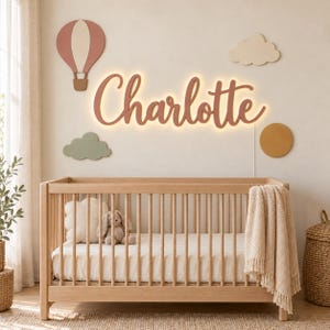 May include: A wooden crib with a light-colored finish, a white mattress, and a beige textured blanket. Above the crib, a wall-mounted sign displays the name Charlotte in cursive script with warm backlighting, surrounded by wall decor including a hot air balloon, two clouds, and a circle.