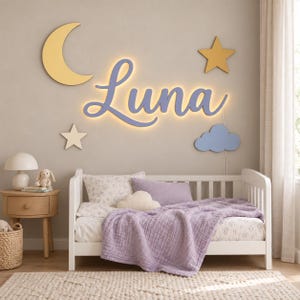 May include: A wall-mounted light fixture featuring the name Luna in purple script lettering with a warm backlight, accompanied by a yellow crescent moon, a yellow star, a white star, and a blue cloud cutout mounted on the wall above a bed.
