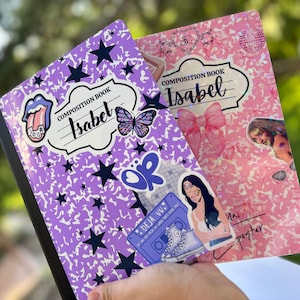 Puede incluir: Dos cuadernos de composición, uno morado y otro rosa, se sostienen en la mano. El cuaderno morado tiene un diseño de estrellas y mariposas con el nombre "Isabel" en la portada. El cuaderno rosa tiene un lazo rosa y un diseño de bola de discoteca con el nombre "Isabel".