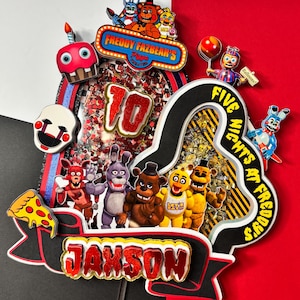 May include: A colorful birthday cake topper featuring characters from "Five Nights at Freddy's." The design includes the text "Freddy Fazbear's Pizza," "Five Nights at Freddy's," and the name "JAYSON." The number "10" is also displayed.