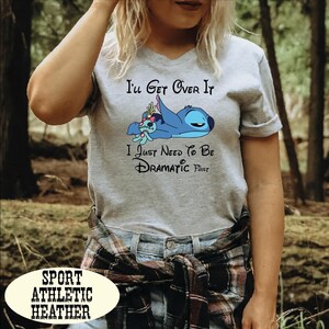 A woman wearing a gray t-shirt with a graphic design of a cartoon character and text that says "I'll Get Over It I Just Need To Be Dramatic First".