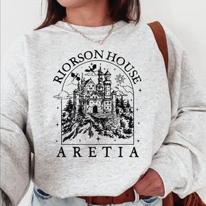 A person wearing a gray sweatshirt with a graphic design depicting a castle and the text "Rierson House Areia" printed on it.