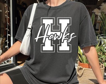 May include: Dark gray t-shirt with the word "Hawks" in white script over a white block letter "H". The shirt is worn with black shorts and a white shoulder bag. The shirt appears to be made of a soft, comfortable material.