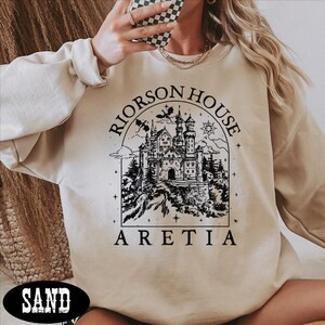 A person wearing a beige sweater with a graphic design of a castle and the text "Riorson House Aretia" printed on it.