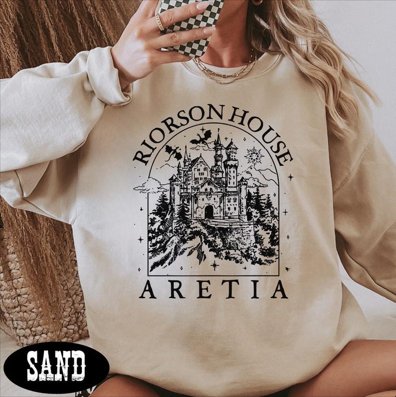 A person wearing a beige sweater with a graphic design of a castle and the text "Riorson House Aretia" printed on it.