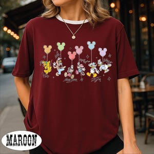 A woman wearing a burgundy t-shirt with a Disney character design, including Mickey Mouse, Donald Duck, and Goofy, along with balloons.