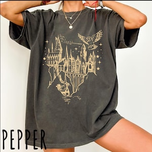 May include: Dark gray t-shirt featuring a gold illustration of a castle, an owl, and stars. The castle is set on a cliff with water below. The word "PEPPER" is printed in white at the bottom left.