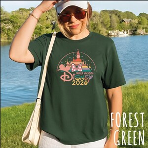 A woman wearing a green t-shirt with a Disney-themed graphic and sunglasses stands in front of a lake with trees in the background.