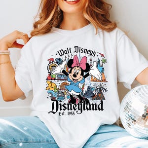 A woman wearing a white t-shirt with a Disney-themed graphic and text, including Minnie Mouse and various Disney landmarks.