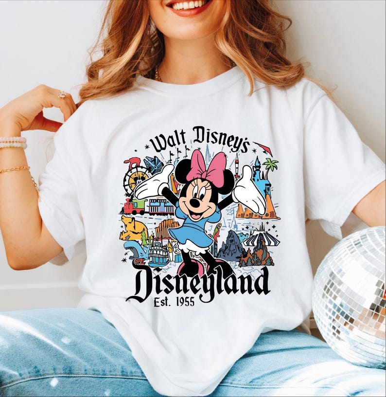 A woman wearing a white t-shirt with a Disney-themed graphic and text, including Minnie Mouse and various Disney landmarks.