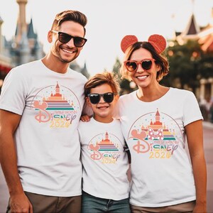 A family of three, consisting of a man, a woman, and a child, is standing together and smiling, wearing matching Disney-themed t-shirts.
