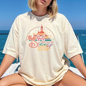 A woman wearing a white t-shirt with a Disney-themed graphic and text, sitting on a boat in front of a blue ocean.