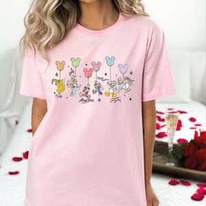 A woman wearing a pink t-shirt with a Disney character design, including Mickey Mouse, Donald Duck, Goofy, and Pluto, along with colorful balloons.