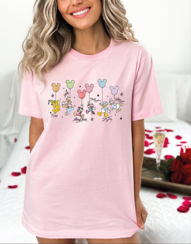 A woman wearing a pink t-shirt with a Disney character design, including Mickey Mouse, Donald Duck, Goofy, and Pluto, along with colorful balloons.