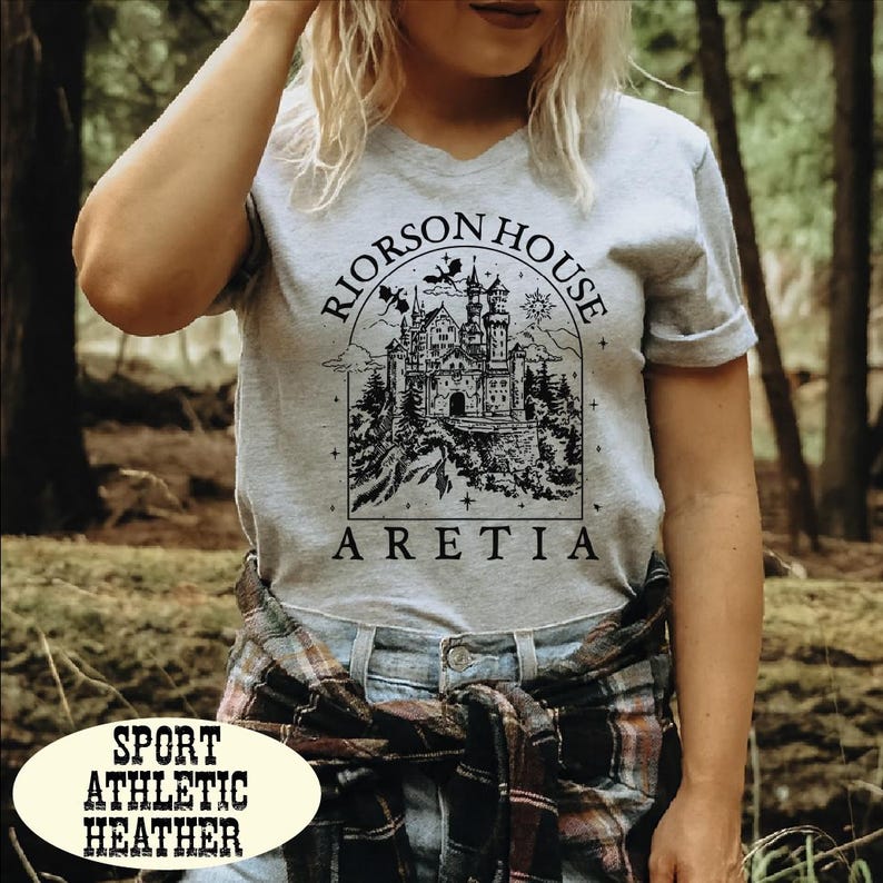A woman wearing a grey t-shirt with a graphic design of a castle and the text "Riorson House Aretia" printed on it, standing in a forested area.