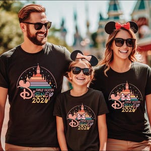 A family of three, consisting of a man, a woman, and a child, are standing together and smiling, all wearing matching Disney-themed t-shirts.