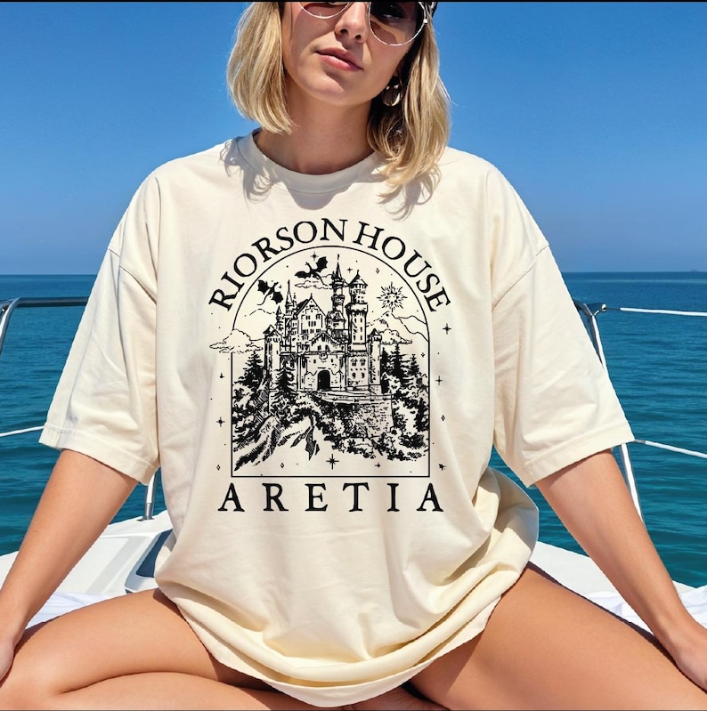 A woman wearing a white t-shirt with a graphic design of a castle and the text "Rierson House Areia" is sitting on a boat in the ocean.