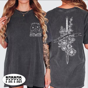 May include: Dark gray t-shirt with white graphic designs. The front features a mountain and book design with text. The back displays a sword, flowers, and the words "You do not yield". The shirt has a vintage, worn look.