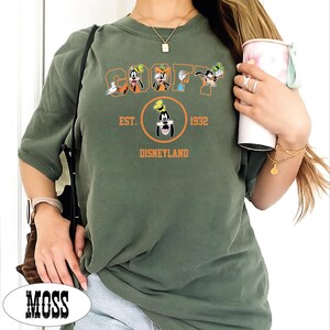 A person wearing a green t-shirt with a graphic of Goofy and Mickey Mouse, holding a cup, and a necklace.