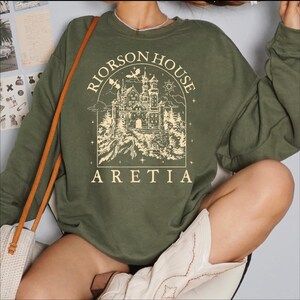 A person wearing a green sweatshirt with a graphic design of a castle and the text "Rierson House Areia" printed on it.
