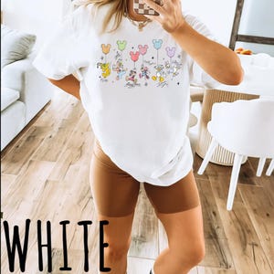 A person is taking a selfie in a room with a white t-shirt featuring a graphic of Disney characters and balloons.