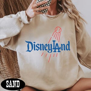 A person wearing a beige sweatshirt with the text "Disneyland" printed on it, holding a phone up to their face.