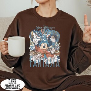 A woman wearing a brown sweatshirt with a Disney character graphic and the text "FANTASIA" printed on it, holding a white mug.