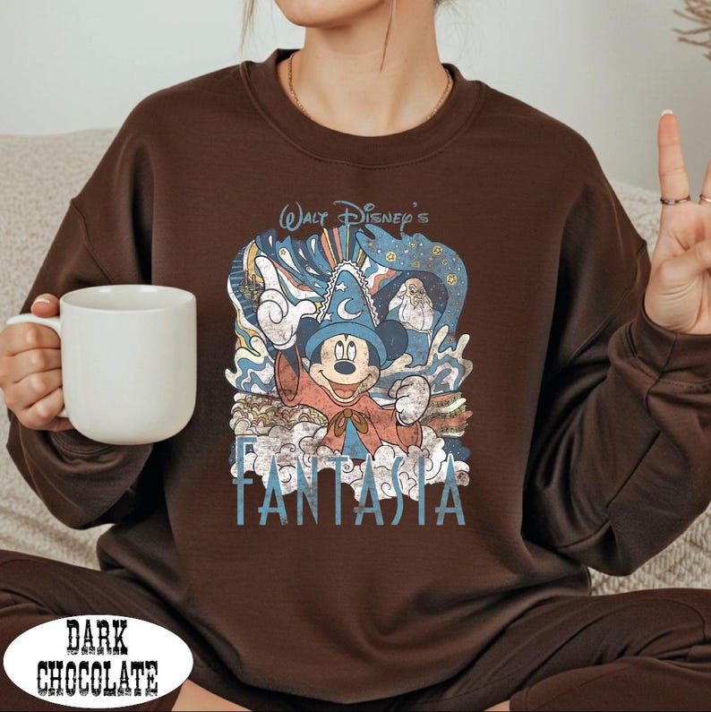 A woman wearing a brown sweatshirt with a Disney character graphic and the text "FANTASIA" printed on it, holding a white mug.