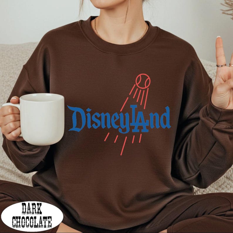 A person sitting on a couch wearing a brown sweater with the word "Disneyland" printed on it, holding a white mug and making a peace sign gesture.
