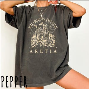 A person wearing a gray t-shirt with a graphic design of a castle and the text "Riorson House Aretia" printed on it.