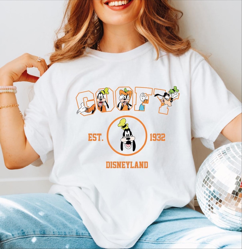 A woman wearing a white t-shirt with a graphic design featuring Goofy and Mickey Mouse characters, along with text that says "Est. 1932 DISNEYLAND".