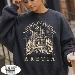 A woman wearing a gray sweatshirt with a graphic design of a castle and the text "RIORSON HOUSE ARETIA" printed on it.