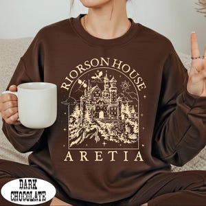 A person wearing a brown sweater with a graphic design of a castle and the text "Rierson House Aretia" is sitting on a couch and holding a white mug.