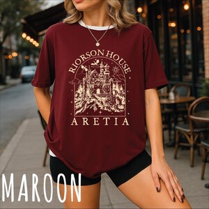 A woman wearing a burgundy t-shirt with a graphic design of a castle and the text "Riorson House Aretia" printed on it, standing on a sidewalk.