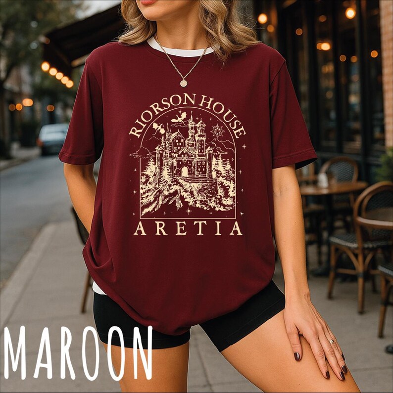 A woman wearing a burgundy t-shirt with a graphic design of a castle and the text "Riorson House Aretia" printed on it, standing on a sidewalk.