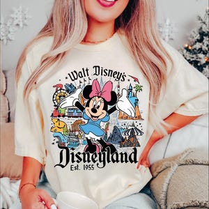 A woman wearing a white t-shirt with a Disney-themed graphic and text, smiling and holding a cup.