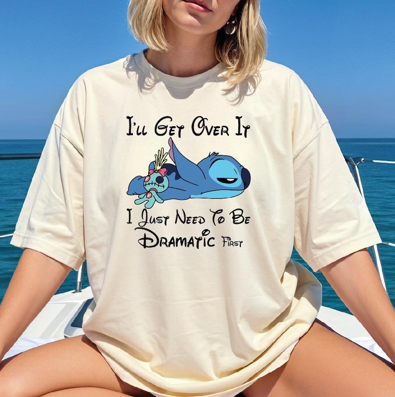 A person wearing a white t-shirt with a graphic design of a cartoon character and text that says "I'll Get Over It I Just Need To Be Dramatic First".