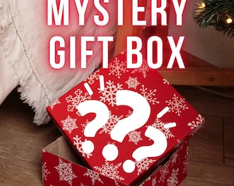 Puede incluir: Una caja de regalo roja con copos de nieve blancos y dos grandes signos de interrogación blancos en la tapa. Las palabras "MYSTERY GIFT BOX" se muestran en letras blancas grandes y brillantes. La caja está abierta.