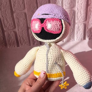 Op de afbeelding: Handgemaakte gehaakte pop met een ronde kop, met een lavendelkleurige pet en een roze bril. De pop heeft een crèmekleurig lichaam met een geel en grijs geaccentueerd jasje. Een kleine gele ster hangt aan het jasje.