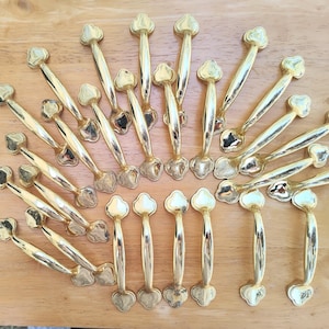 May include: A collection of gold-colored cabinet pulls with a decorative floral design. The pulls are arranged on a light brown wooden surface. The pulls have a curved handle and a decorative base.