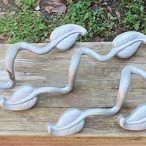 May include: Four silver leaf-shaped hooks with a vine design. The hooks are attached to a wooden surface.