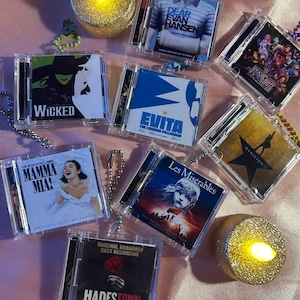Może przedstawiać: Miniaturowe etui na płyty CD z produkcjami musicalowymi, takimi jak "Wicked", "Evita" i "Dear Evan Hansen". Każde etui to replika płyty CD, z tytułem i grafiką przedstawienia. Etui są przymocowane do breloków.