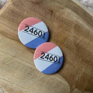 Può includere: Due spille rotonde con uno schema di colori rosso, bianco e blu. Ogni spilla mostra il numero nero "24601". Le spille sono su una superficie di legno.
