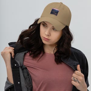 May include: A tan baseball cap featuring a square graphic on the front panel. The graphic depicts a winding road through a dark mountain landscape under a pink sky, with the text "RUN FAR LIVE FREE" printed in small letters beneath the image.