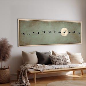 Peut inclure: Une œuvre d'art murale rectangulaire panoramique représentant un vol d'oiseaux noirs sur un fond vert sauge atténué, avec une grande pleine lune jaune pâle située sur le côté droit, le tout encadré par un cadre fin en bois clair.