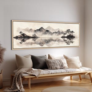 May include: A framed landscape art print depicts a serene mountain scene with a lake. The artwork features a muted color palette of black, gray, and beige, with reflections in the water. The print is displayed above a wooden bench with pillows and a blanket.