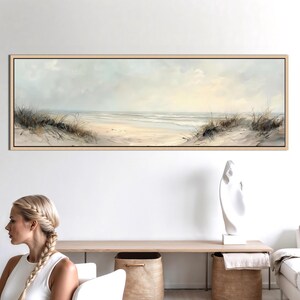 May include: A panoramic framed wall art print depicting a coastal beach scene with soft beige sand, gentle ocean waves, and patches of dry dune grass under a pale, overcast sky. The artwork features muted tones of cream, light gray, and tan.