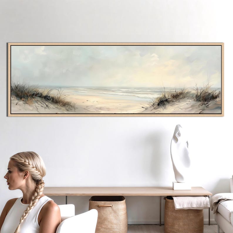 May include: A panoramic framed wall art print depicting a coastal beach scene with soft beige sand, gentle ocean waves, and patches of dry dune grass under a pale, overcast sky. The artwork features muted tones of cream, light gray, and tan.