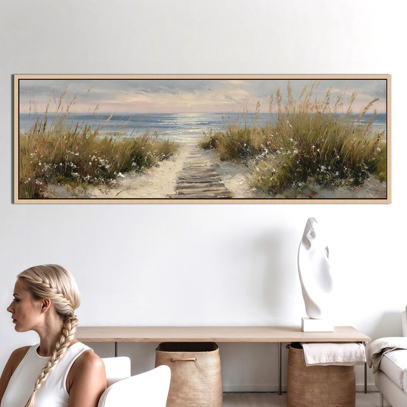 May include: A panoramic landscape painting in a light wood frame depicting a sandy beach path leading to the ocean. The scene features tall dune grasses, soft white flowers, and a calm sea under a muted sky, capturing a serene coastal atmosphere.